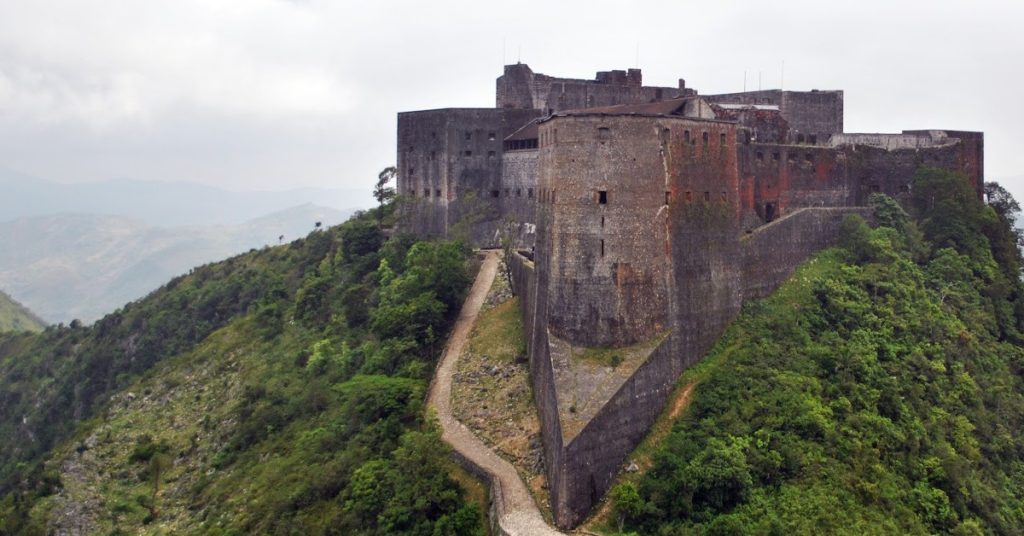 Medieval fortress on hilltop surrounded by lush greenery and mountain landscape, historic castle architecture, Sam Desmet photography.
