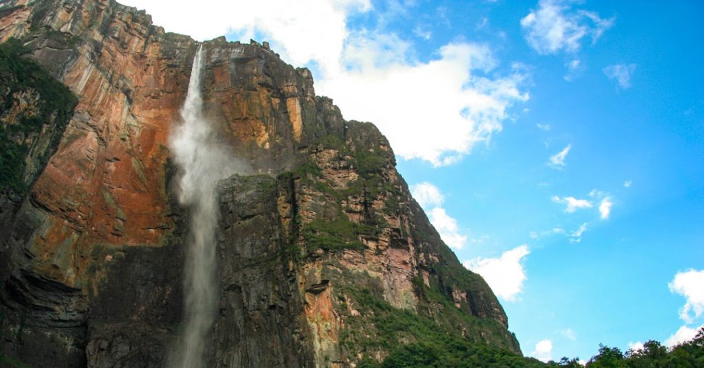 Aerial view of a majestic waterfall cascading down a rugged mountain cliff under a bright blue sky with scattered clouds.