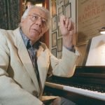 Elderly man playing piano in a cozy room with framed posters on the wall.