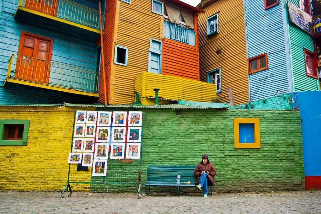 Colorful urban building facade with street art, vibrant painted walls, and a person sitting on a bench in an artistic neighborhood.