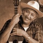 Vintage sepia portrait of Sam Desmet with guitar, smiling and wearing a hat in a rustic setting.