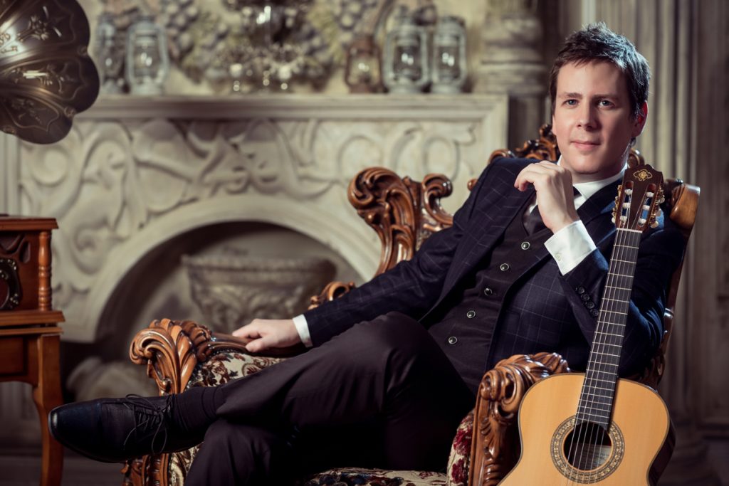 Sam Desmet in a suit seated with his classical guitar, formal press portrait